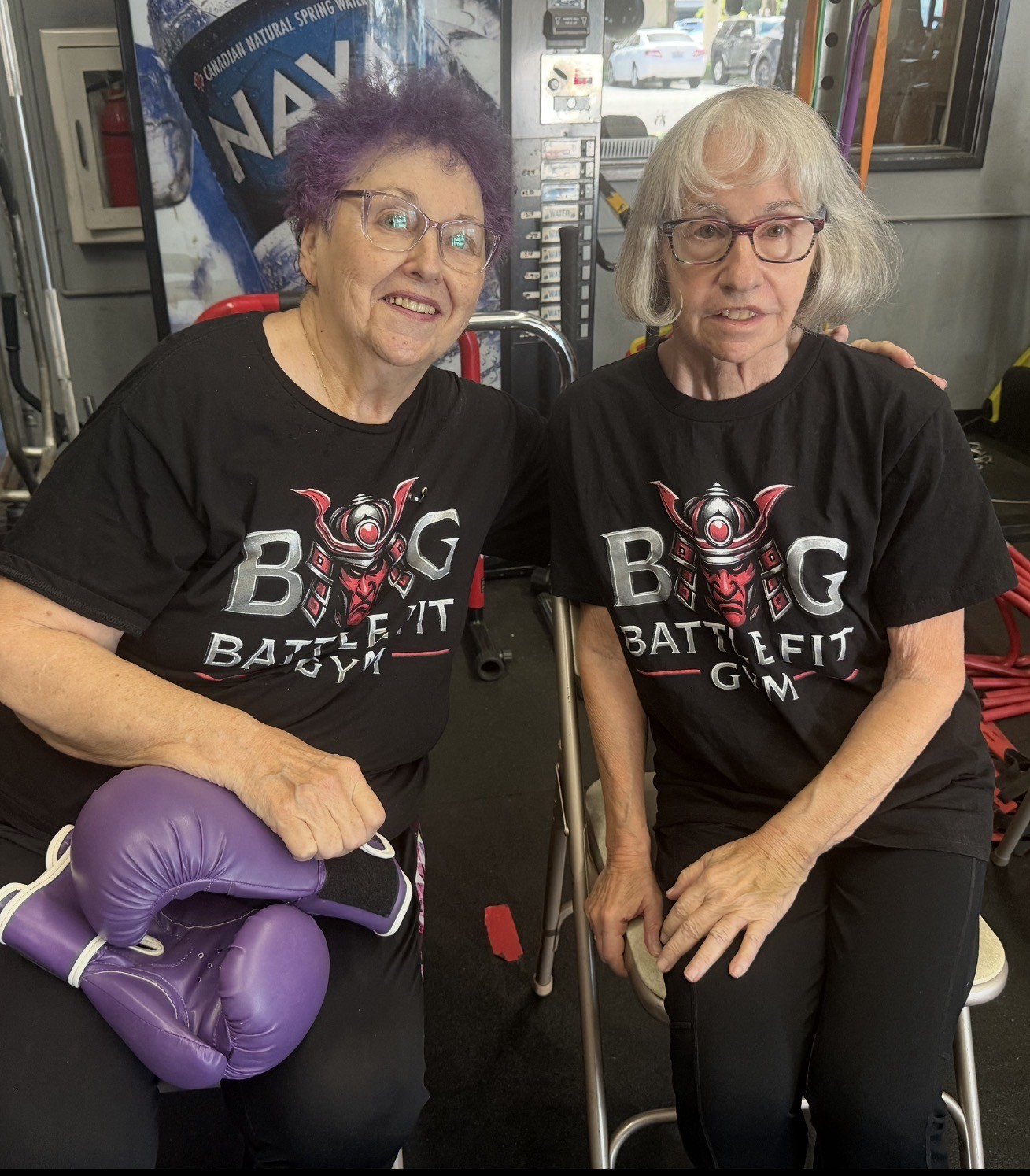 Two smiling senior members wearing Battlefit Gym shirts, with boxing gloves